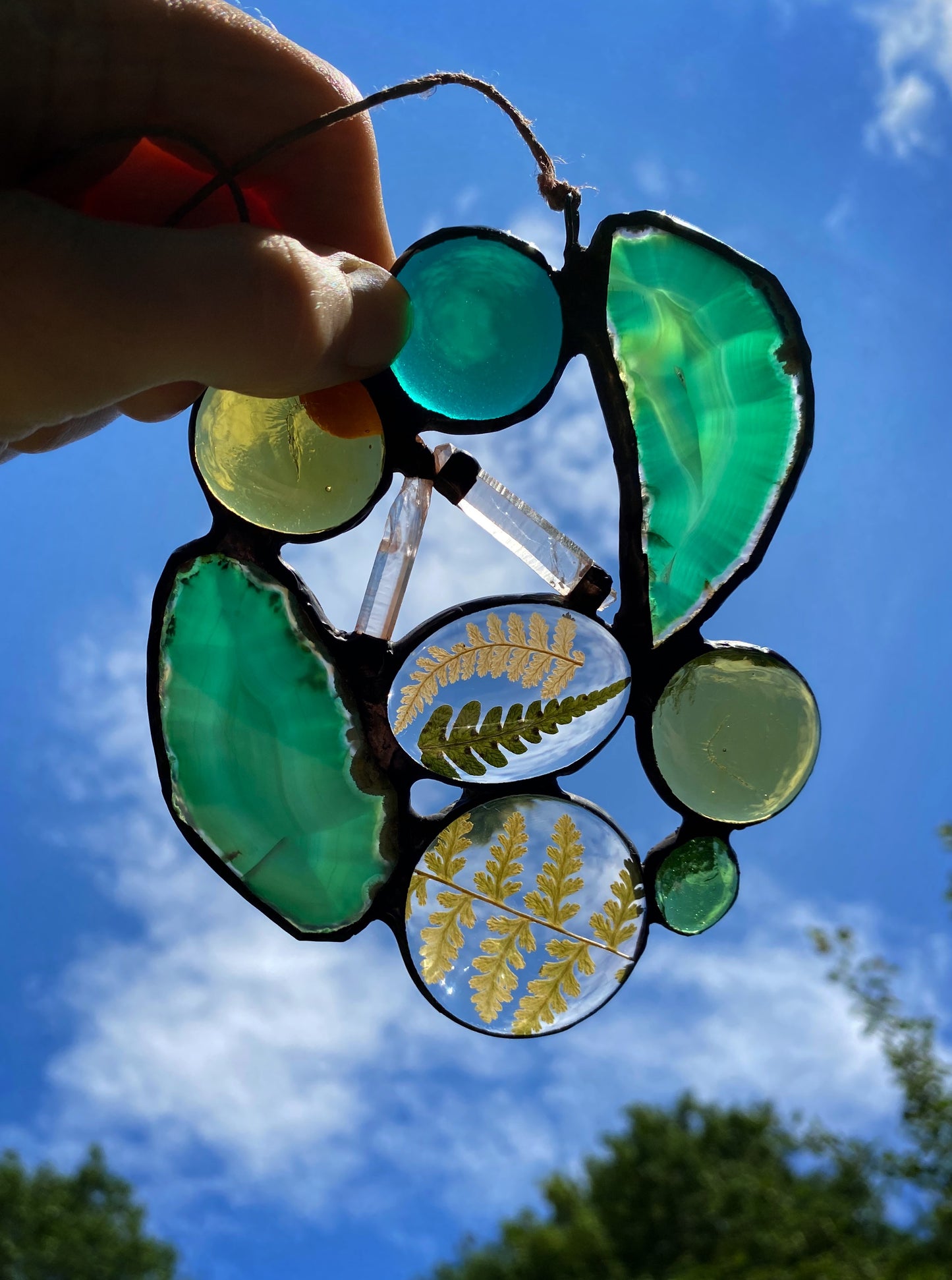 Glass art suncatcher with agate slices quartz crystals and ferns against a blue sky.