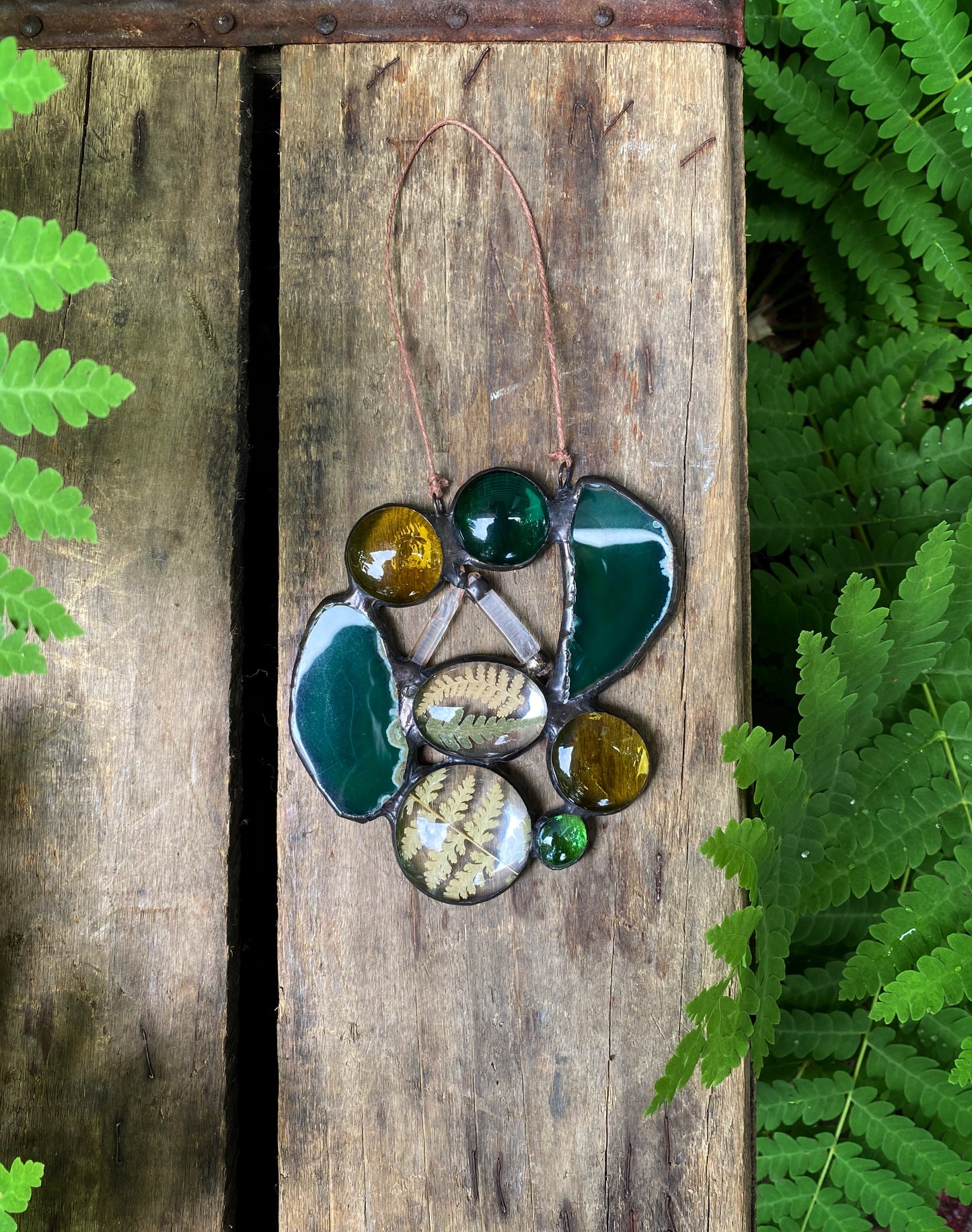 Stained glass suncatcher with quartz crystals pressed ferns and agate slices on a wooden board with ferns in the background.