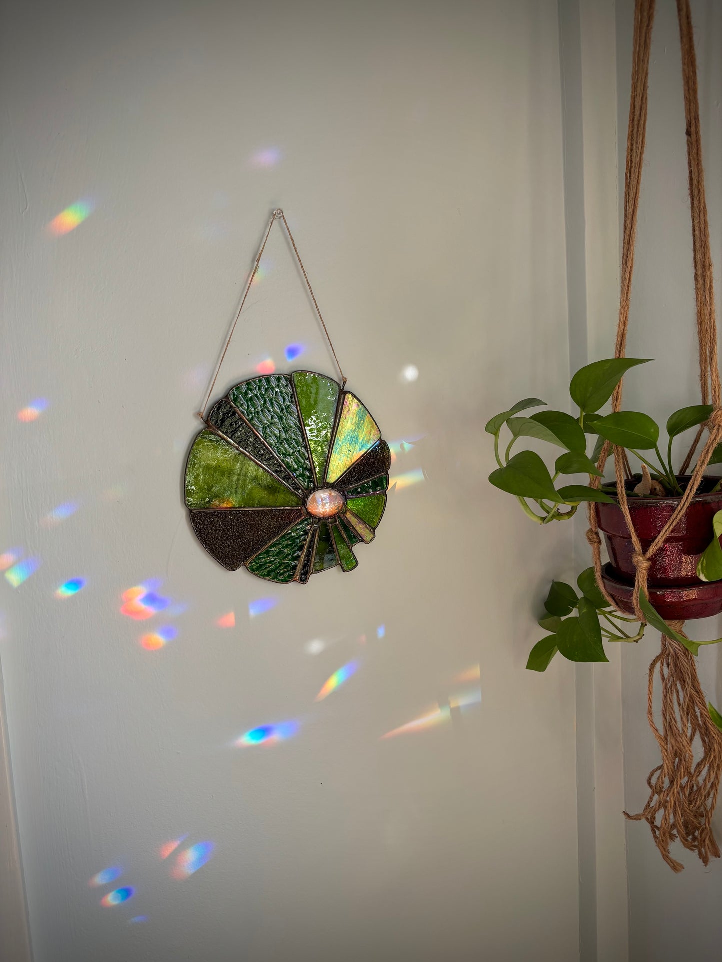 Decorative stained glass with ferns hanging on a white wall with a plant to the right.
