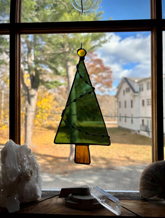 Stained glass Christmas tree hanging on a window with a view of trees and a house outside.