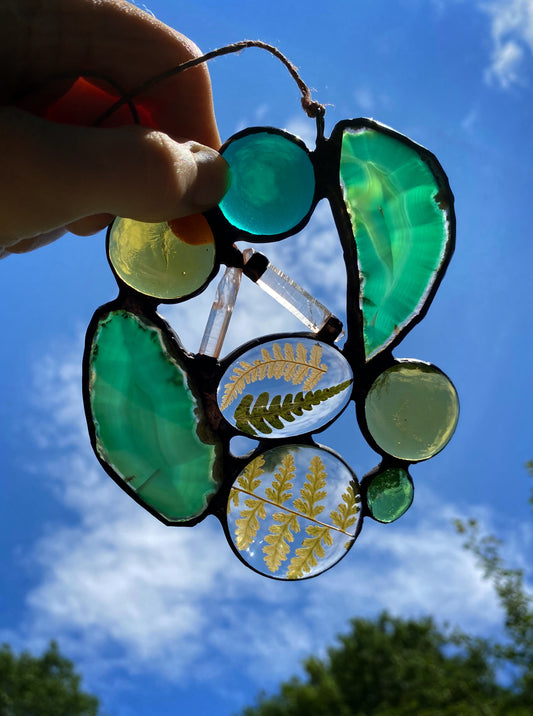 Glass art suncatcher with agate slices quartz crystals and ferns against a blue sky.
