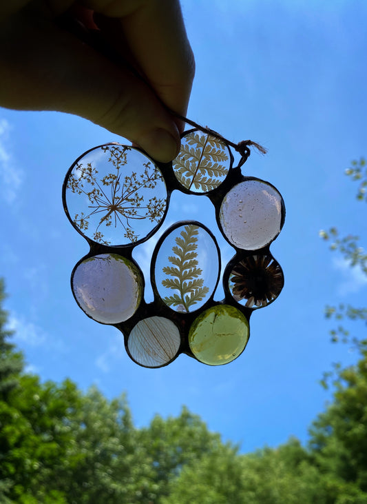 Decorative glass suncatcher with natural elements flowers ferns held against a blue sky.