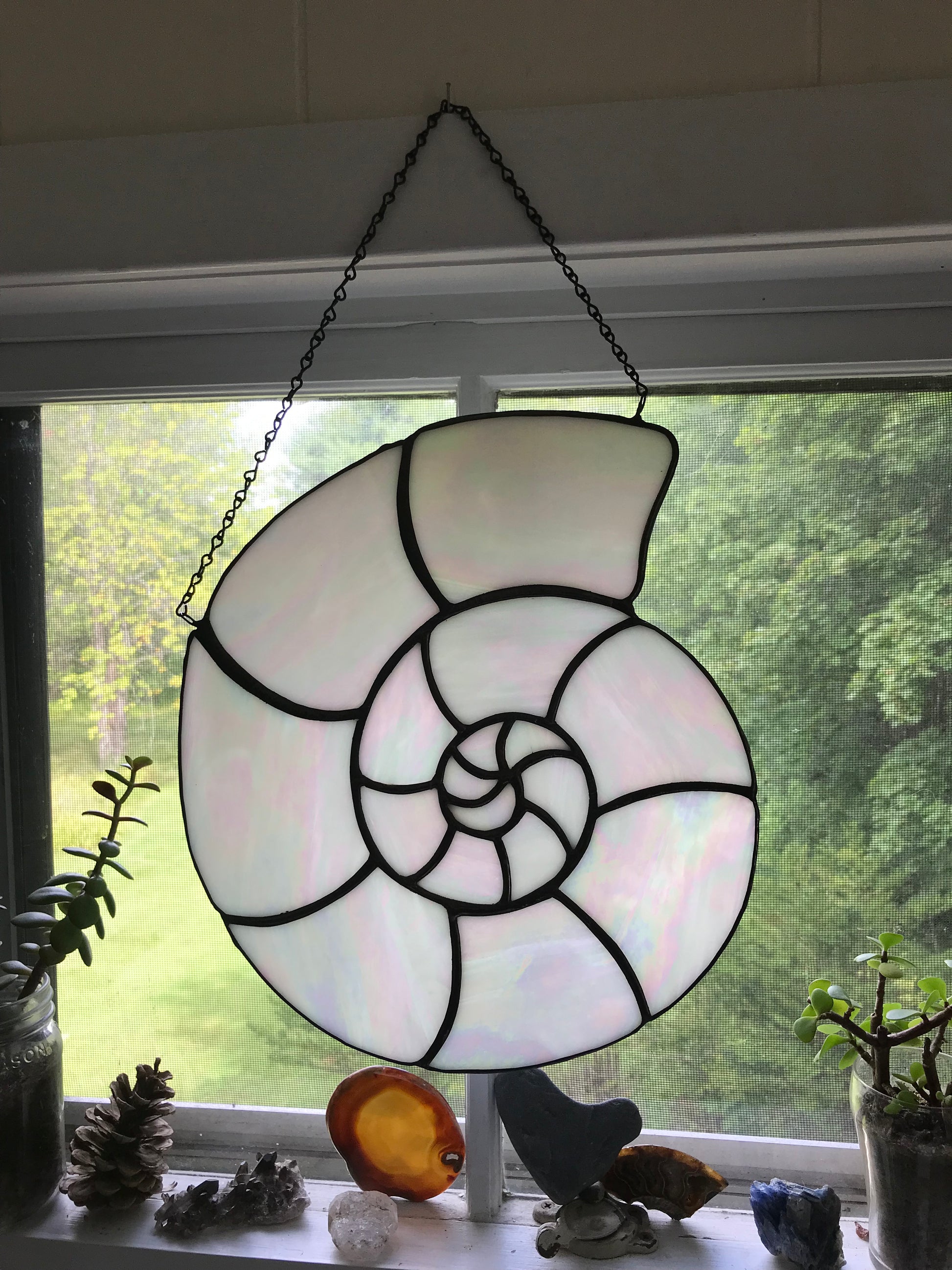 Stained glass nautilus shell hanging in front of a window with greenery outside.