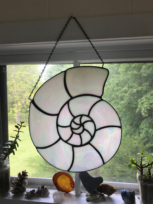 Stained glass nautilus shell hanging in front of a window with greenery outside.