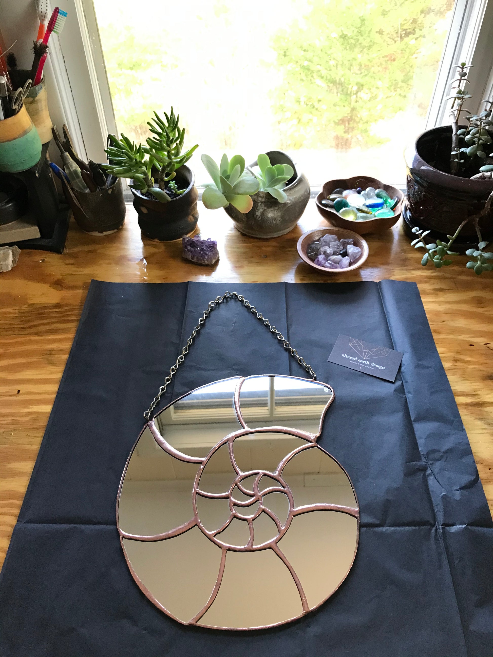 Decorative mirror with a spiral design on a dark blue fabric, placed on a wooden surface with plants in the background.