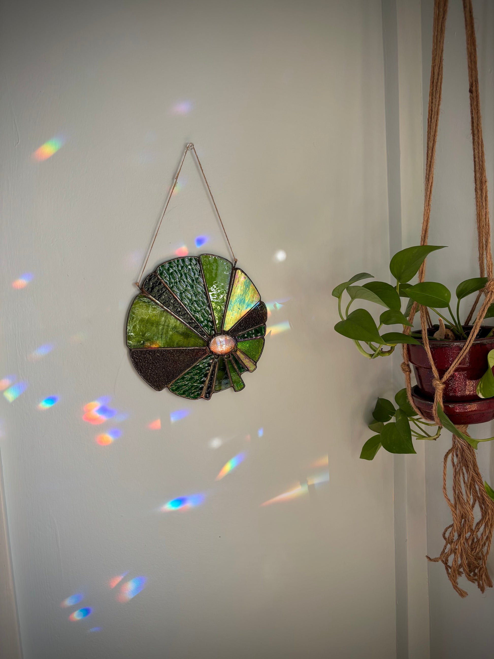 Decorative stained glass with ferns hanging on a white wall with a plant to the right.