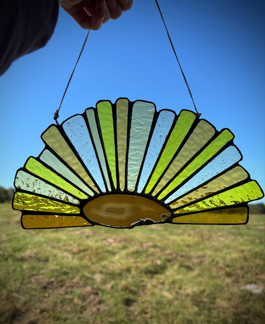 Stained glass sun catcher with an agate slice and fan-like design held against a blue sky.