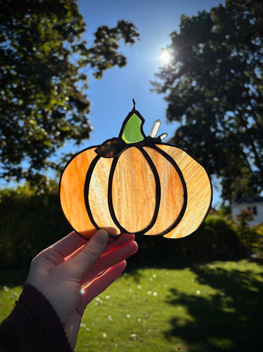 Hand holding a pumpkin-shaped stained glass panel outdoors with trees and sunlight in the background.